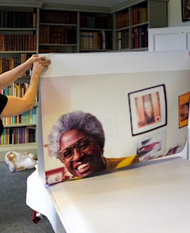 Two hands holding a Folder Poster with an image of a middle age smiling woman of colour wearing glasses. Preparing for the display