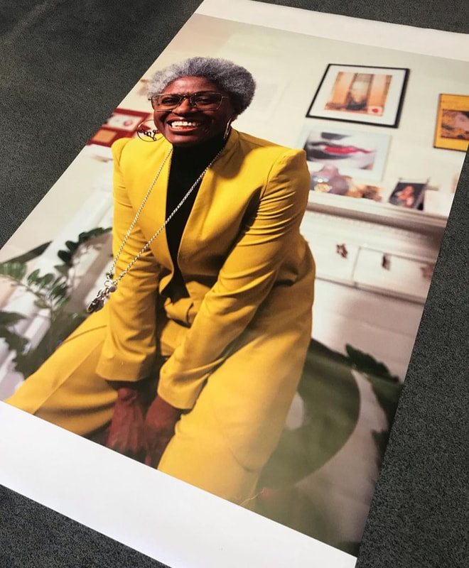 Poster with an image of a middle age smiling woman of colour wearing glasses and a yellow suit. In the background a white fireplace and some plants